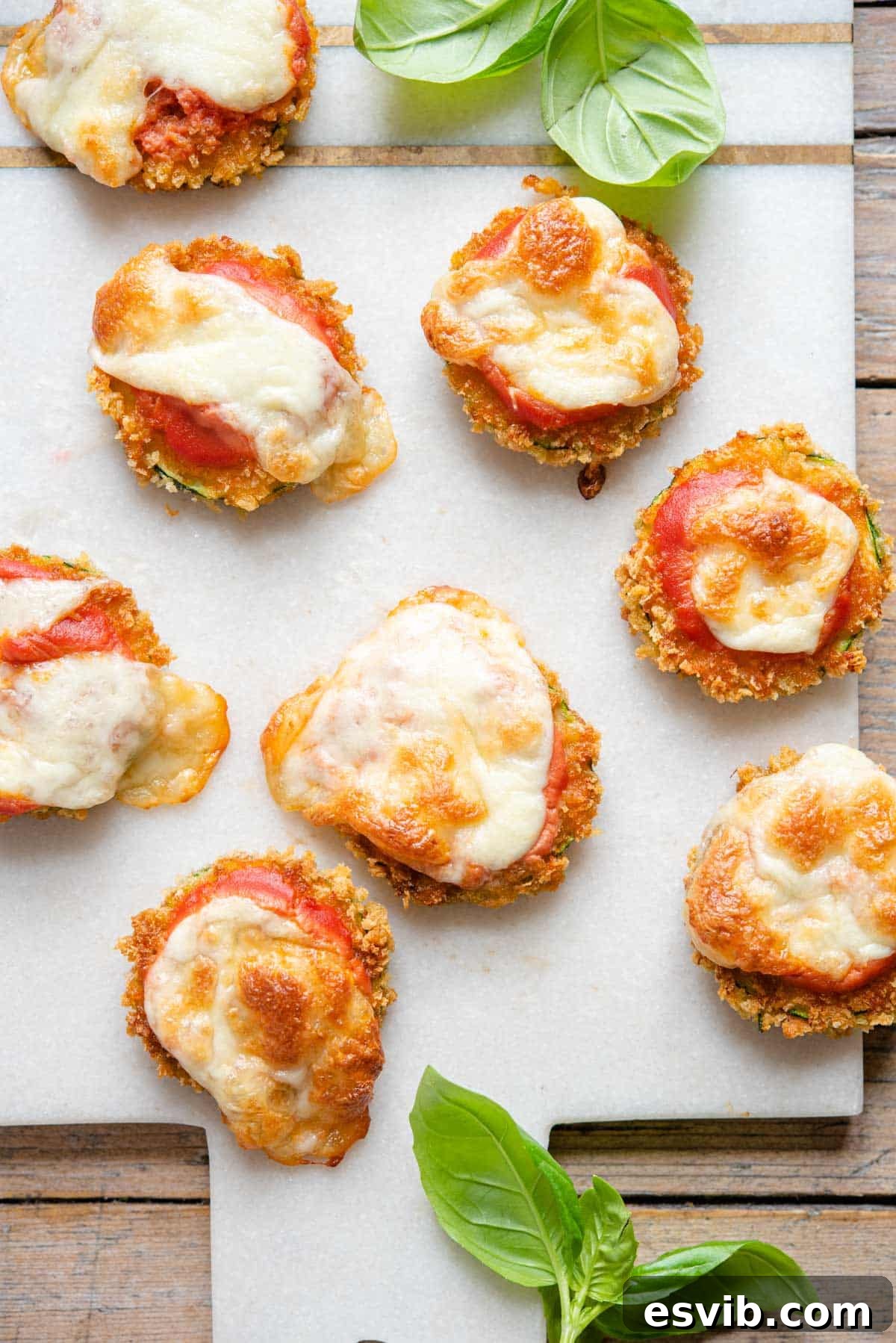 An overhead shot of golden-brown zucchini pizza bites generously topped with melted mozzarella and tomato sauce, arranged on a marble cutting board, ready to be served as a party appetizer.