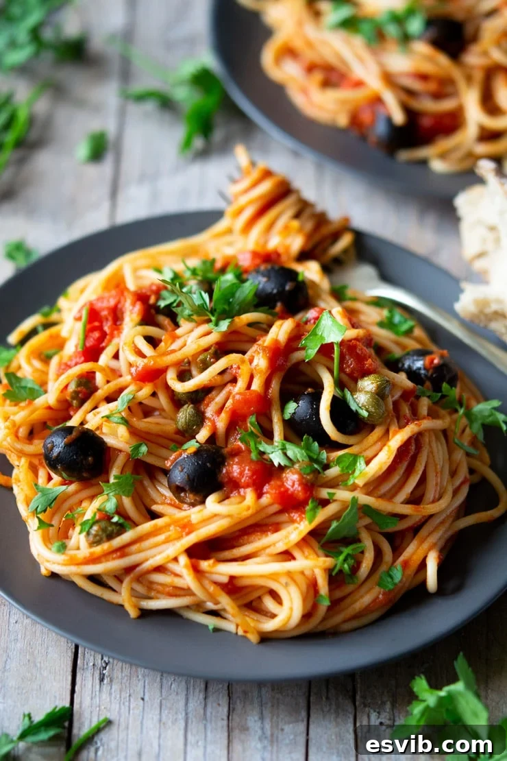 A fantastic Spaghetti alla Puttanesca recipe using fresh cherry tomatoes, capers, olives and garlic it's packed full of flavour and ready in minutes! insidetherustickitchen.com A close up of spaghetti with puttanesca sauce on a white plate, garnished with fresh parsley, sitting on a wooden surface.