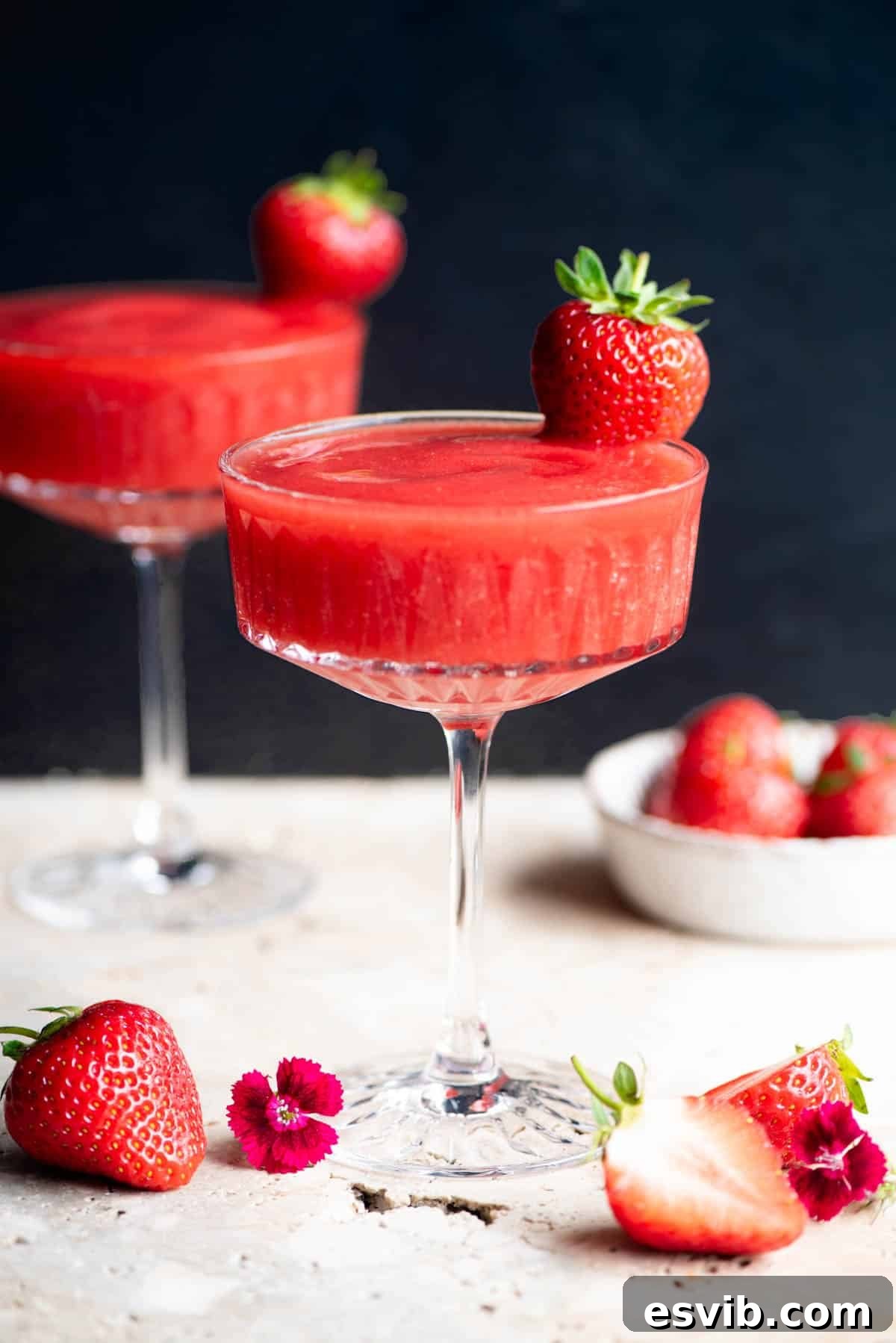Strawberry Velvet Fizz 2 A side shot of a vibrant frozen strawberry cocktail in an elegant stemmed glass, beautifully garnished with a fresh whole strawberry. The background is a sleek black, making the cocktail pop.