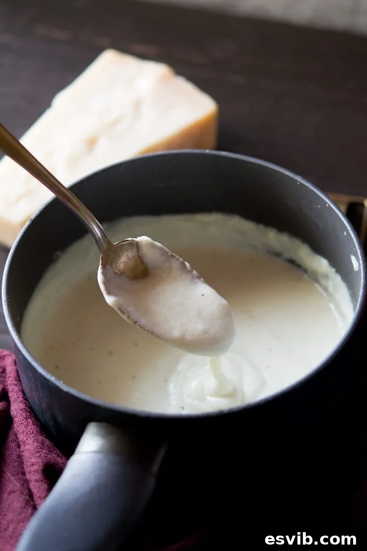 A close-up shot of creamy Bechamel sauce in a saucepan with a bowl of grated Parmesan cheese in the background, ready to be added for a cheese sauce, illustrating the final step.