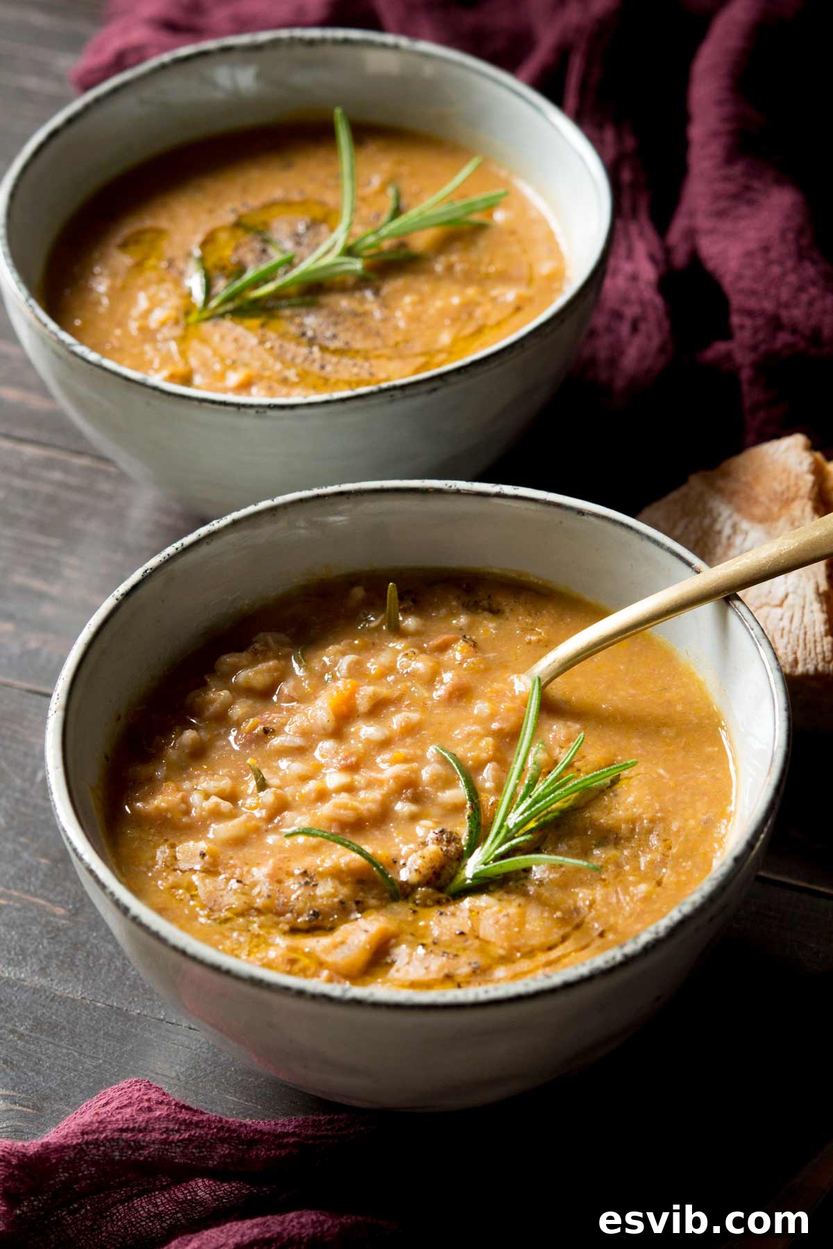 A steaming bowl of rustic Tuscan farro soup, garnished with fresh rosemary, served with a spoon on a wooden surface.