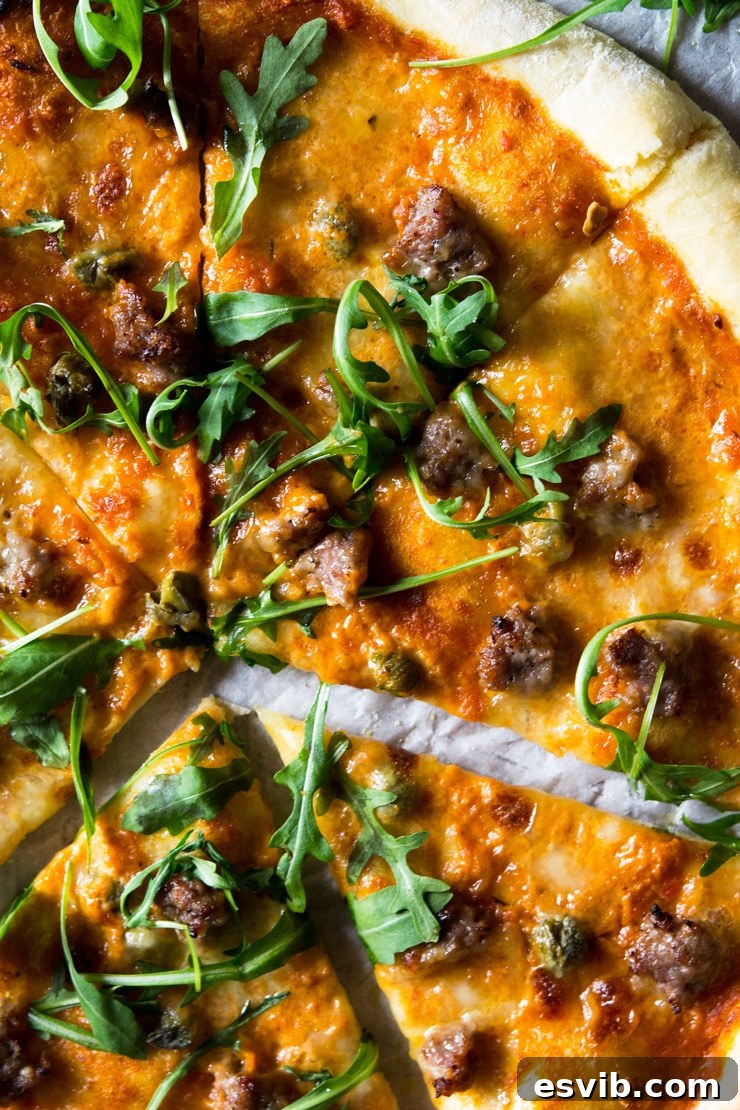 A close-up shot revealing the perfectly melted mozzarella, savory sausage, and fresh arugula on the roasted red pepper pizza. A close up of pizza with sausage and roasted red pepper sauce, melted mozzarella and topped with arugula.