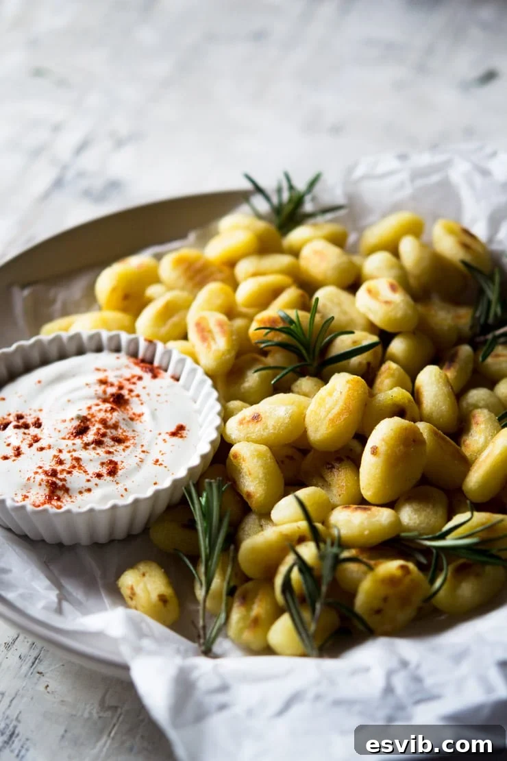 Rosemary Pan-Fried Gnocchi with Dip A close-up shot of crispy pan-fried gnocchi on a white plate, beautifully garnished with fresh rosemary and served alongside a small bowl of sour cream dip