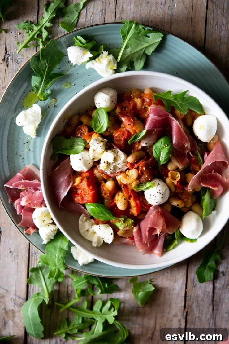 An overhead shot of a vibrant warm white bean salad in a white bowl, generously topped with delicate prosciutto slices and glistening baby mozzarella pearls, ready to be enjoyed.