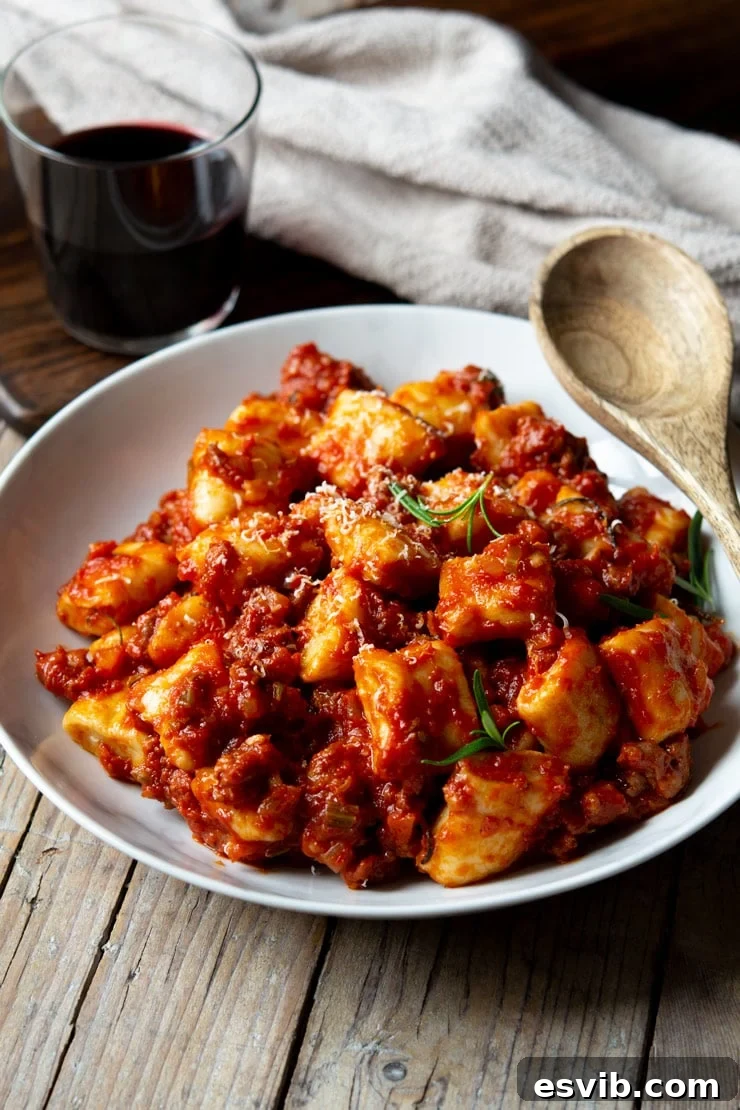 A generous bowl of rich sausage ragu tossed with homemade ricotta gnocchi, with a glass of red wine in the background, evoking cozy Italian comfort.