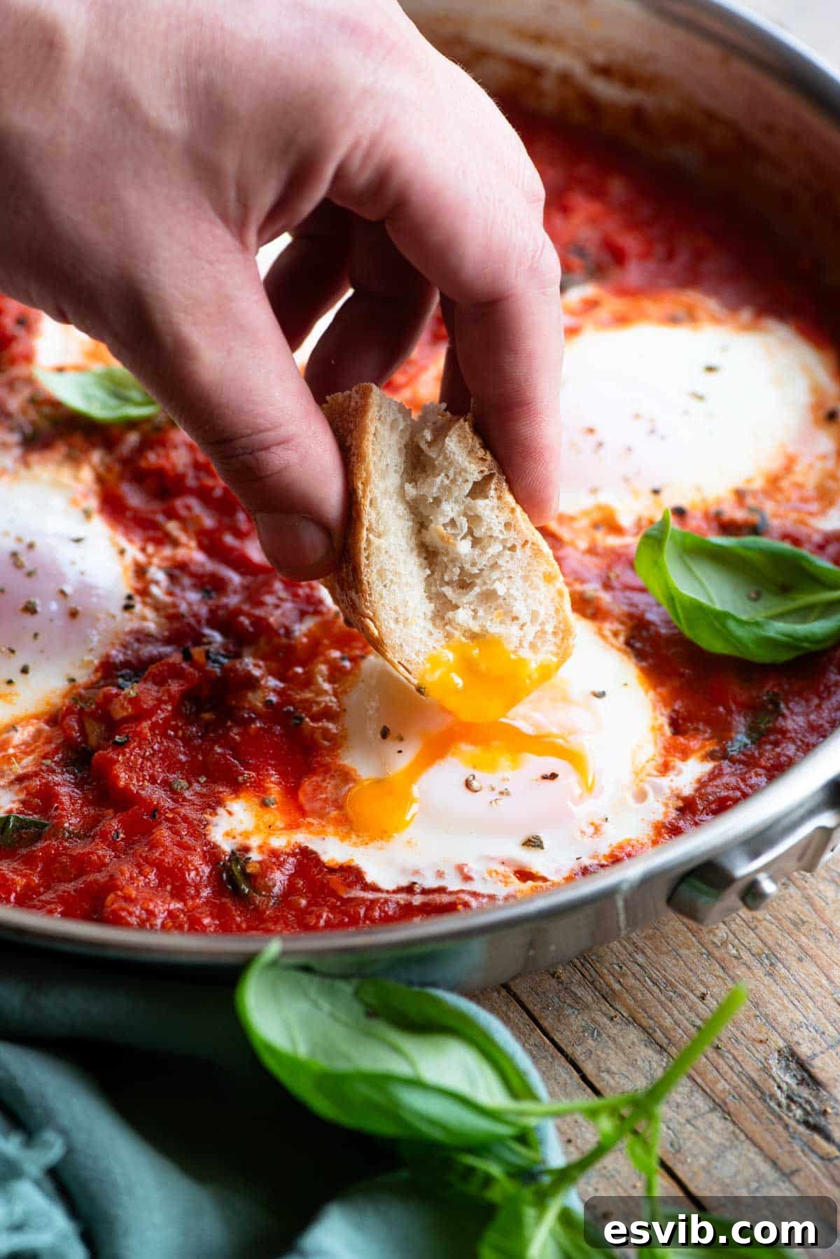 Fiery Nduja Purgatory Eggs 6 A hand dipping some bread into a poached egg with a runny yolk in a pan of tomato sauce.