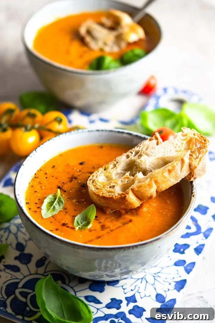 A close up of a bowl of roasted tomato soup with a slice of crusty bread at the side and garnished with basil