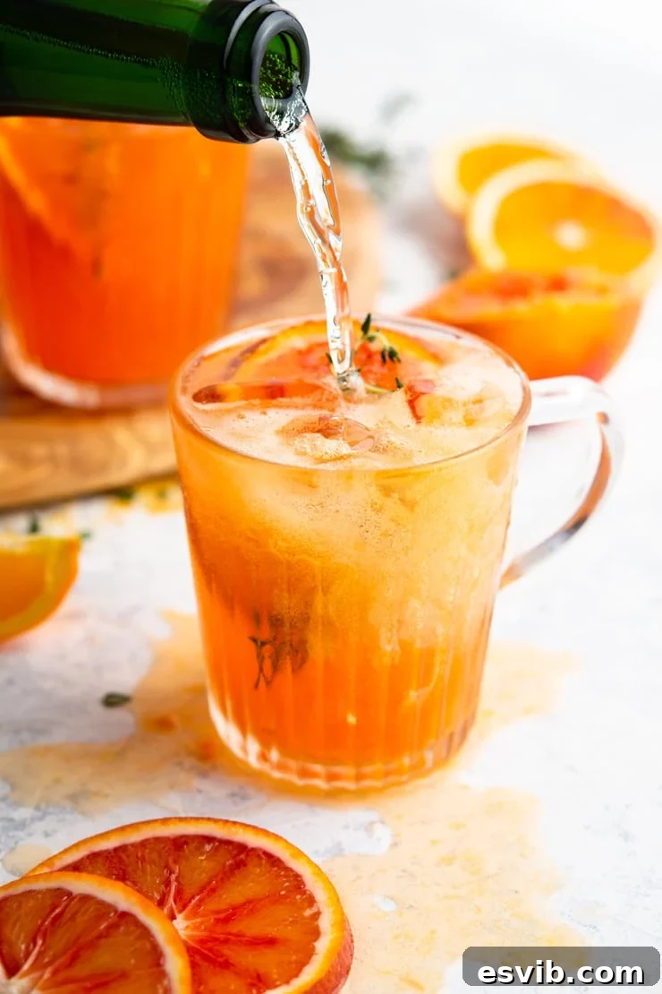 A close-up shot of prosecco being poured into a tall glass containing a blood orange cocktail with ice and a slice of orange, highlighting the effervescence.