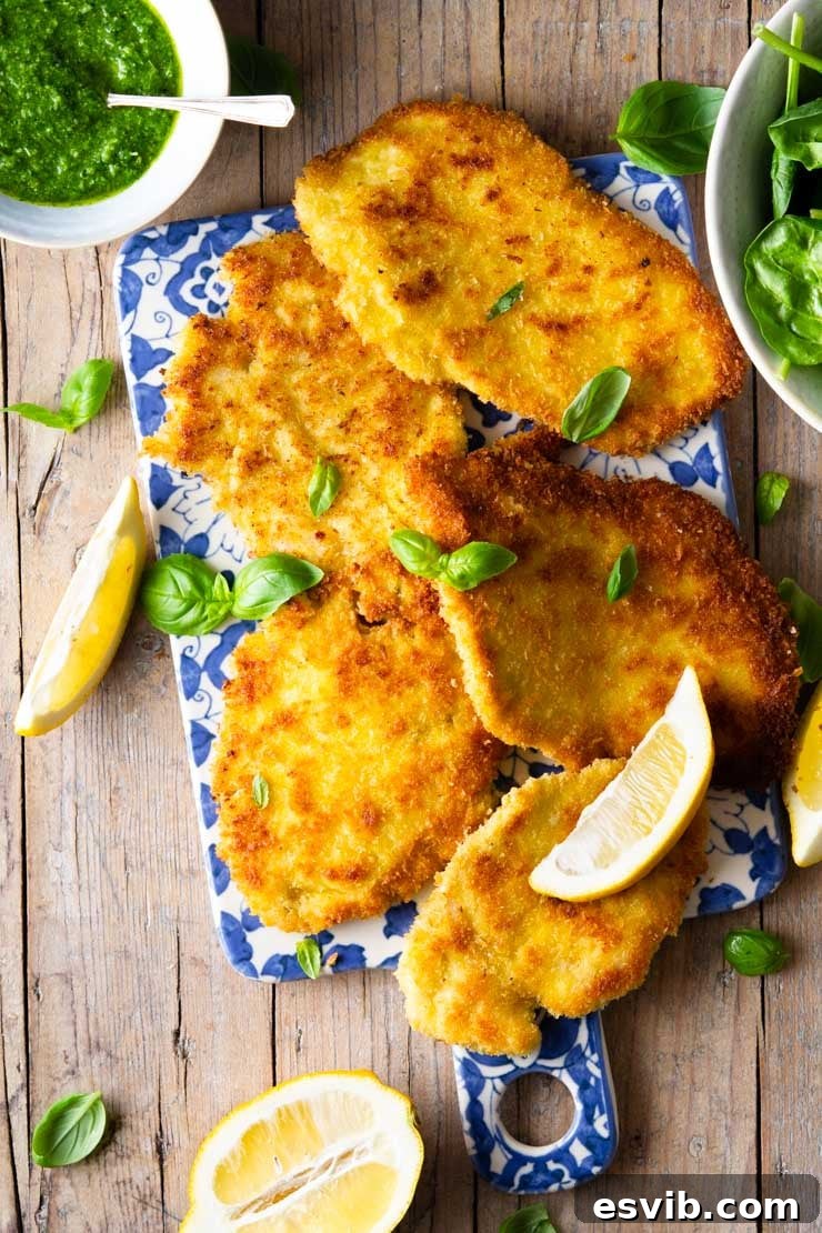 Overhead shot of golden-brown Italian chicken cutlets on a blue board with fresh lemon wedges and a side of green pesto