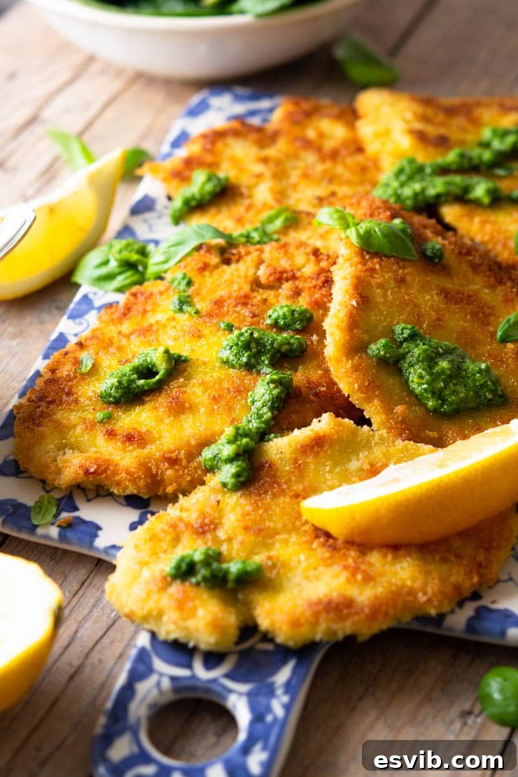 A close-up side shot of a stack of golden-brown breaded chicken cutlets on a blue serving board, garnished with fresh lemon and a drizzle of pesto