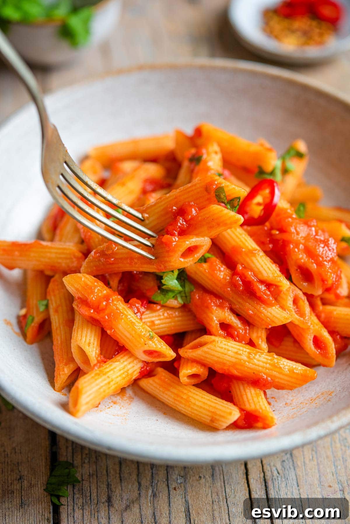 A close up shot of a fork twirling and picking up penne pasta coated in arrabbiata sauce from a white rustic bowl, highlighting the rich tomato color