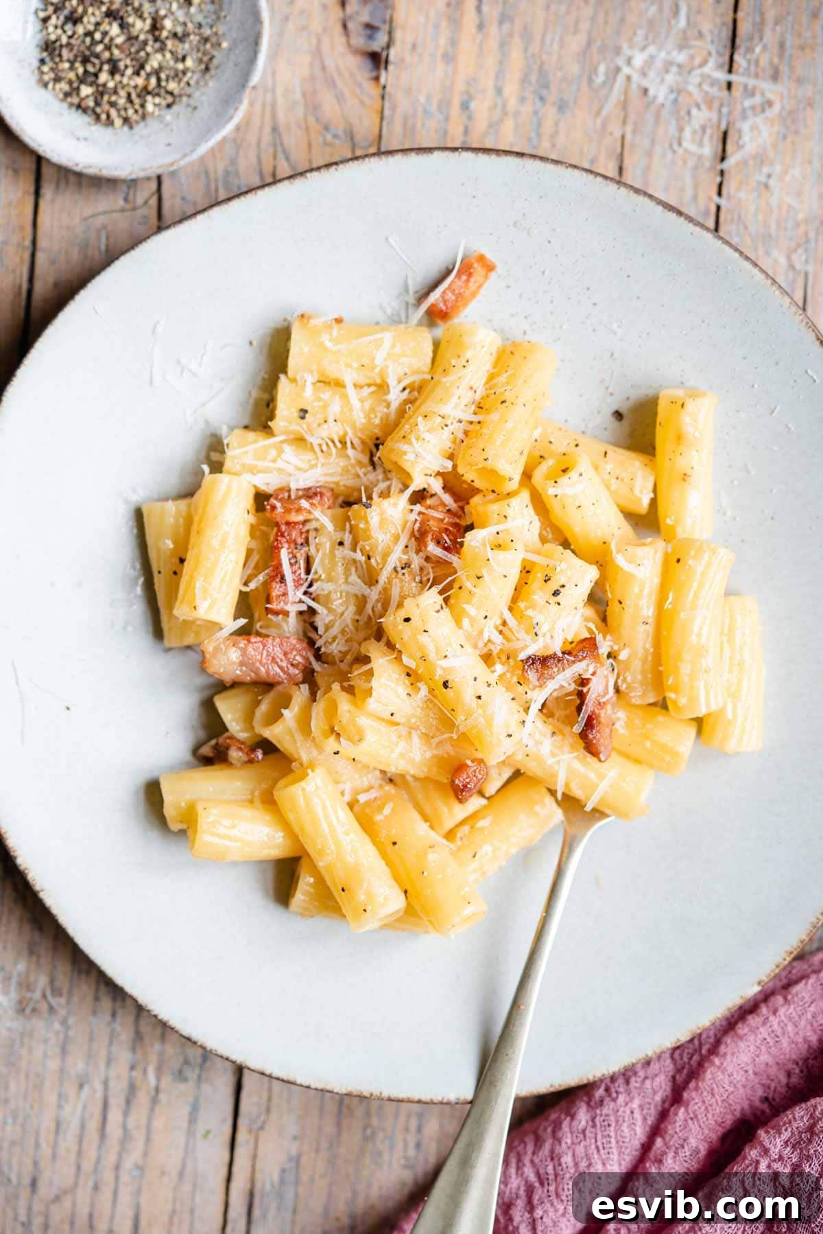 An overhead shot of rigatoni pasta with crispy guanciale on a plate, garnished with fresh black pepper and grated Pecorino Romano