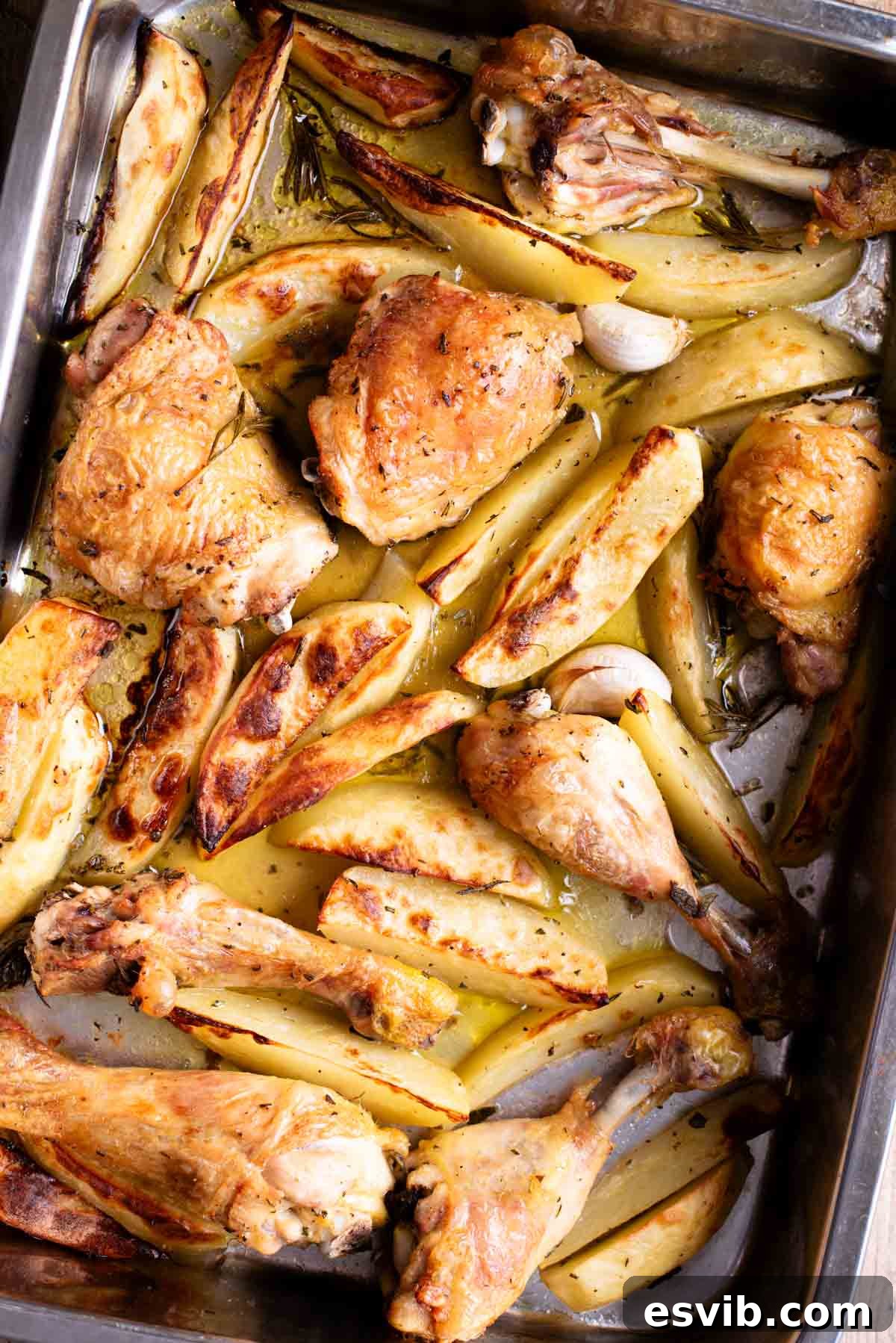 A close up overhead shot of chicken and potato wedges in a roasting tray with garlic and rosemary. Highlighting the golden-brown skin of the chicken and the tender, herb-coated potatoes, ready to be served.