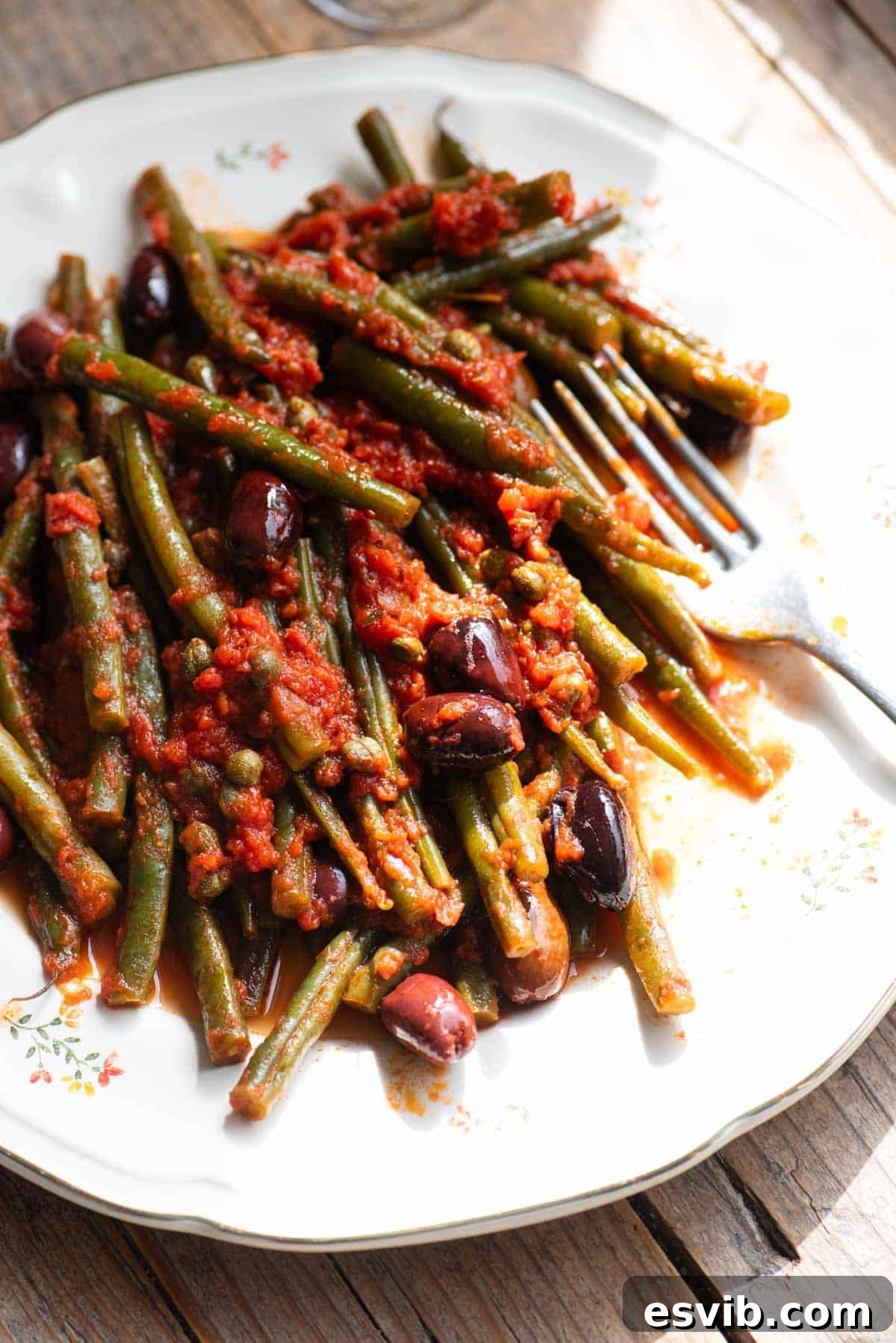 Zesty Green Beans with Olives and Capers 4 A side shot of green beans with tomatoes, olives and capers on a white plate with a fork at the side.