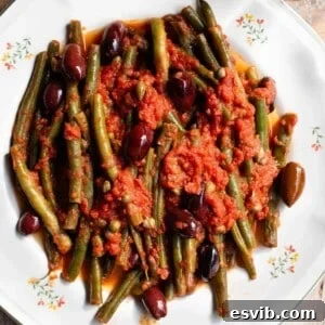 A close up of green beans in tomato sauce with olives and capers on a white plate.