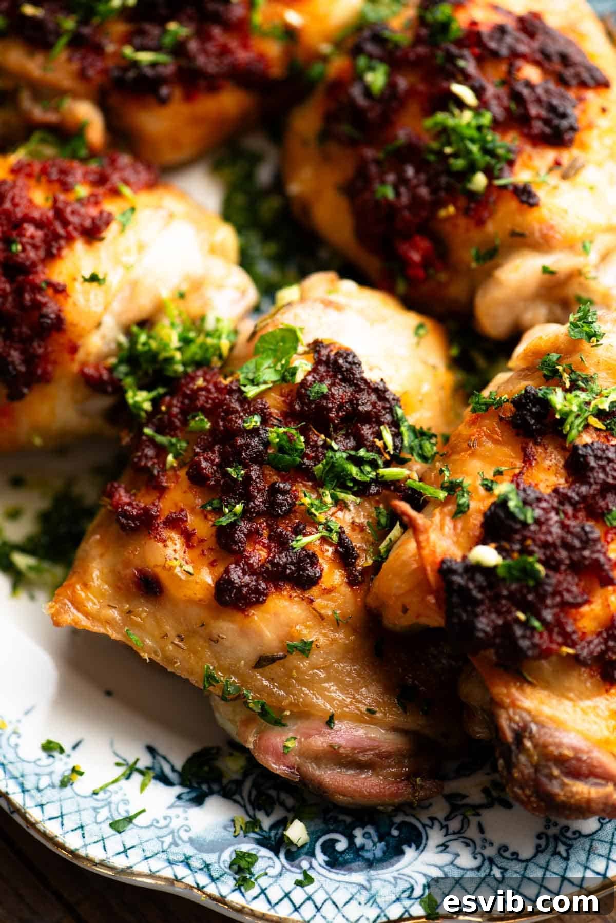 A close-up photograph showcasing the crispy, red-orange skin of a Nduja baked chicken thigh topped with fresh gremolata.