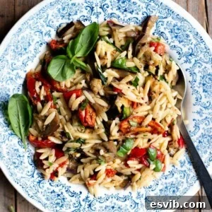 A close up of an orzo pasta salad with grilled veg and tomatoes in a blue bowl with basil on top.