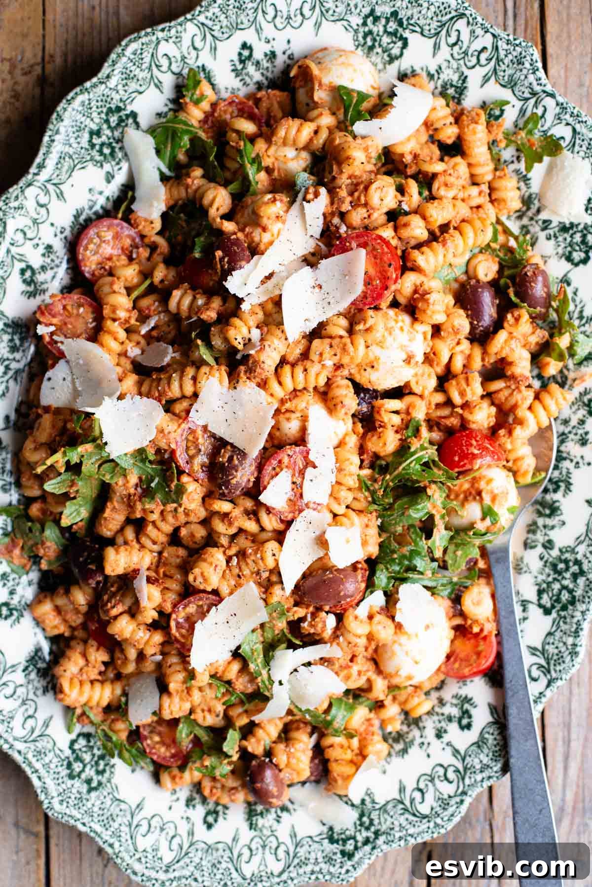 An overhead shot of a sundried tomato pasta salad on a serving plate with mozzarella, olives, arugula and parmigiano shavings.