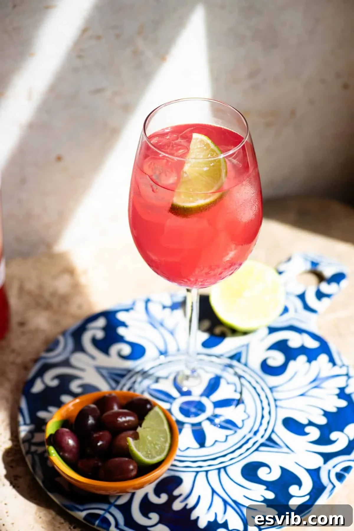 Sarti Spritz Fruity Italian Aperitif Cocktail 4 A close up of a sarti spritz with a wedge of lime on a blue patterned ceramic board with olives at the side.
