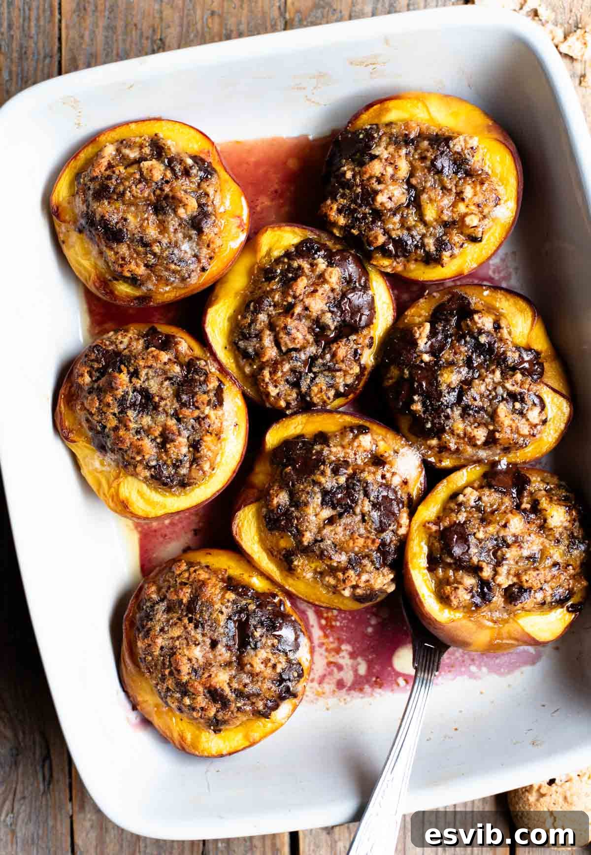 An overhead shot of baked peaches stuffed with amaretti cookies and chocolate in a white baking dish on a rustic wooden background.