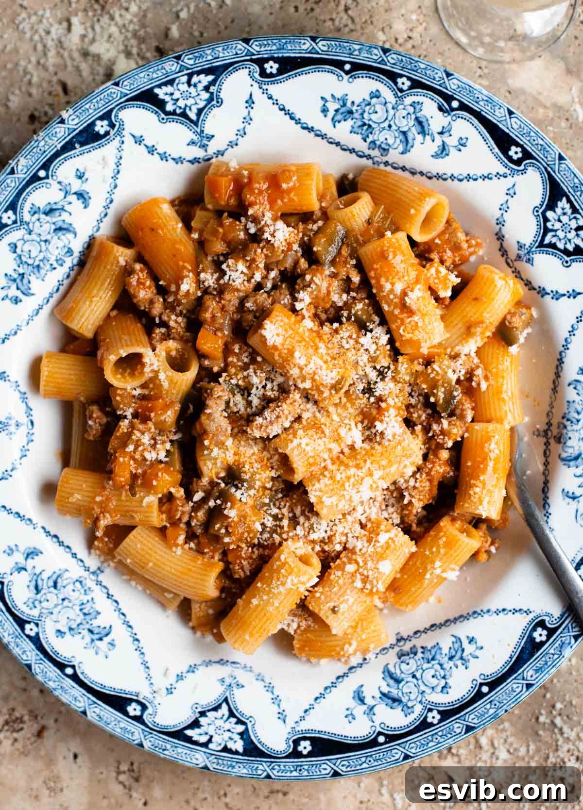 An overhead shot of rigatoni pasta with a sausage and eggplant ragu in a blue and white bowl on a travertine surface.