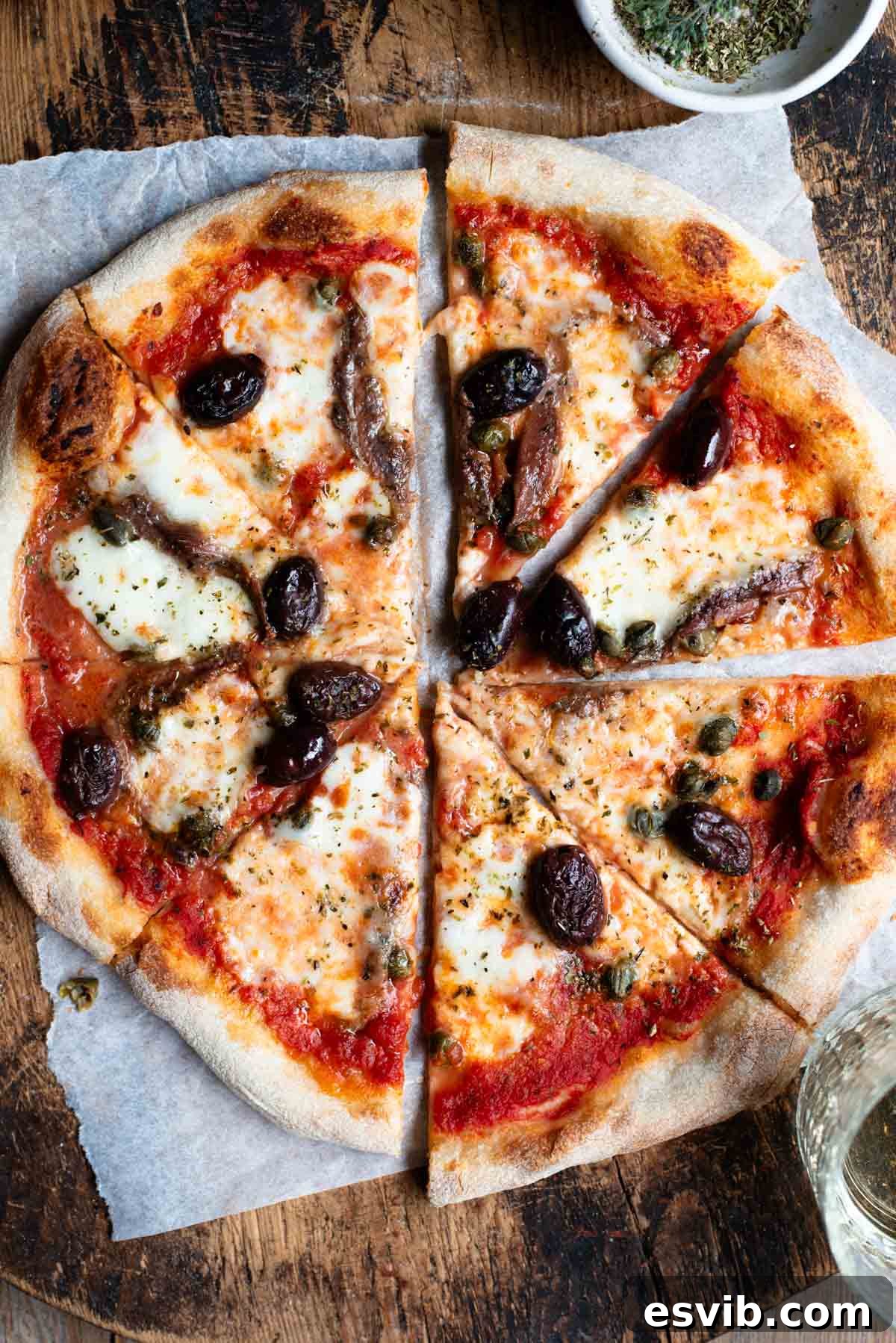 An overhead shot of a pizza cut into slices on a rustic wooden board. The pizza is topped with olives, anchovies and capers.