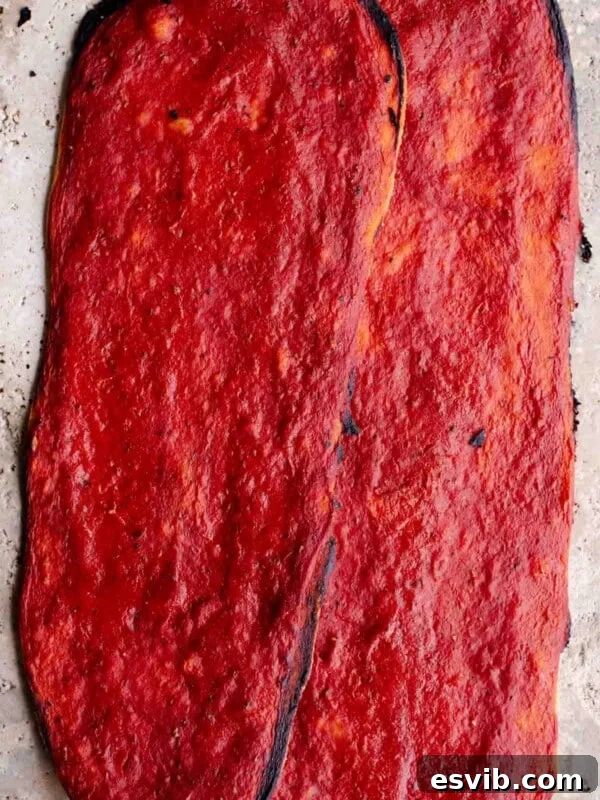 Two long thin and crispy pizzas covered in tomato sauce sitting on a travertine surface.