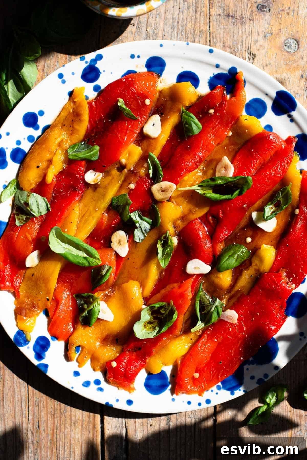 Italian Marinated Roasted Bell Peppers Recipe 9 Red and yellow marinated roasted peppers arranged on a blue speckled plate topped with garlic and basil.