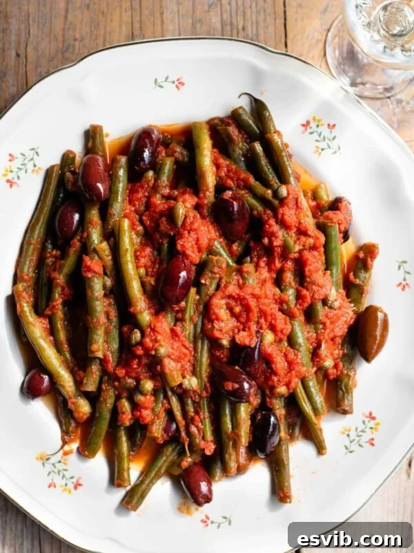 A close up of green beans in tomato sauce with olives and capers on a white floral plate and rustic wooden background.